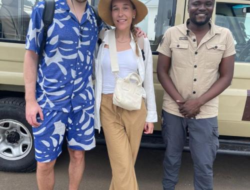 Two excite d visitors and a knowledgeable guide stand on an African savanna, pointing towards a distant elephant herd, amidst golden grasses and acacia trees under a bright blue sky.